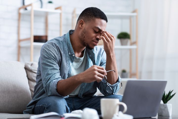 Fatigue oculaire, la reconnaître et la soigner : un jeune homme devant un écran d'ordinateur posé trop bas retire ses lunettes, ferme les yeux et se pince le haut du nez, visiblement fatigué.